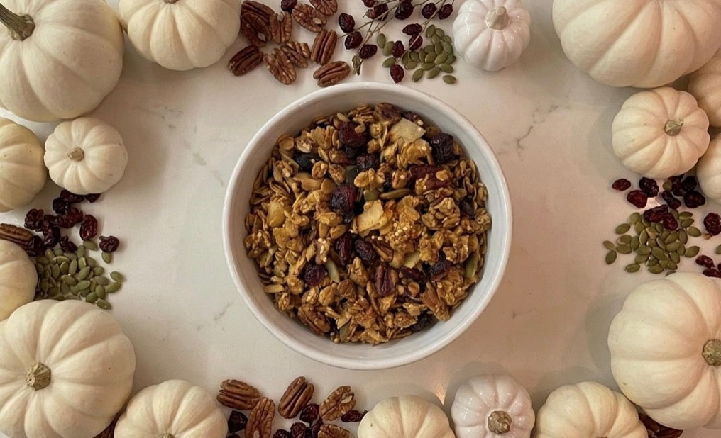 Bowl of Angie's pumpkin spice granola surrounded by white pumpkins, pecans, and cranberries